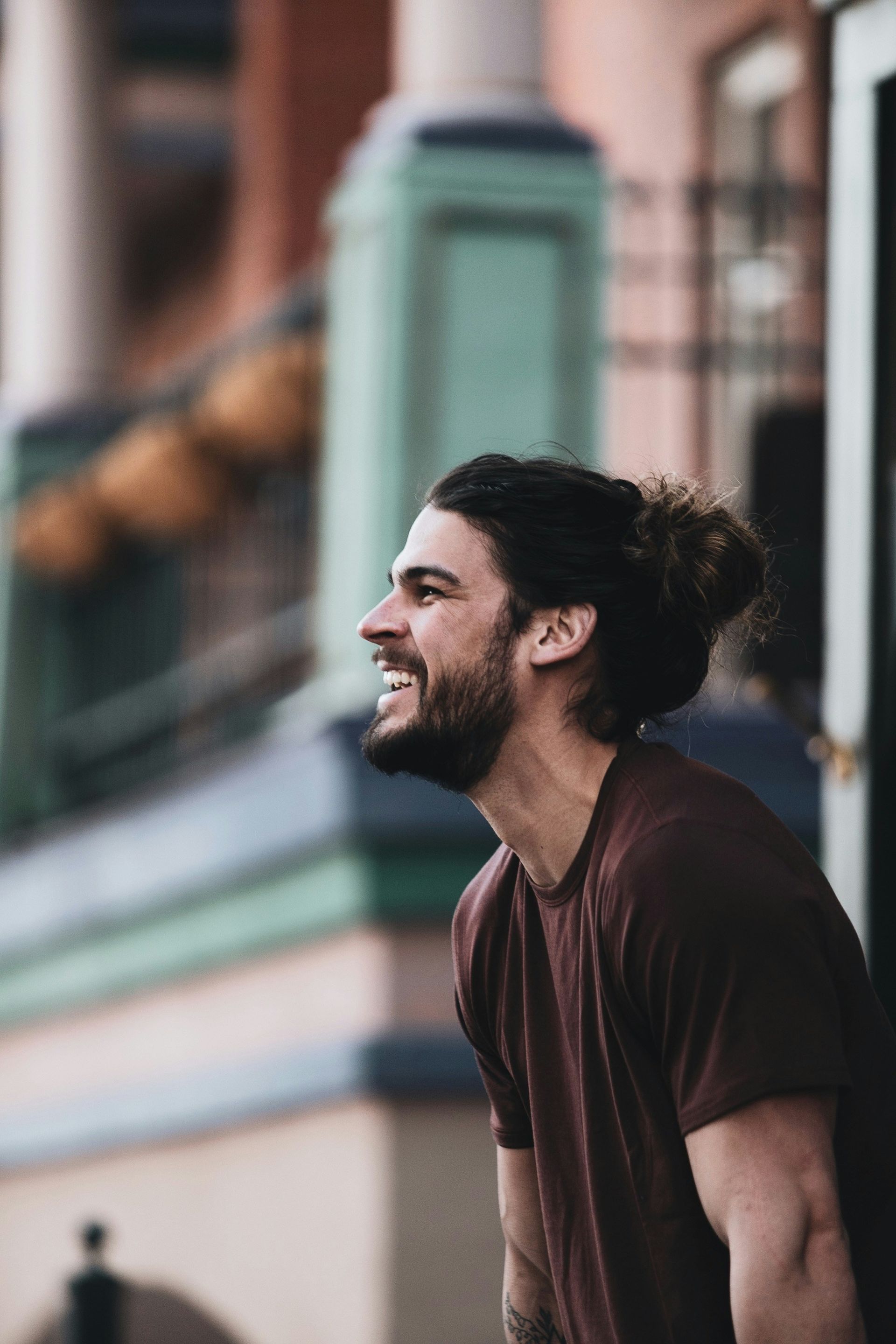 Man with dark hair in a bun, smiling widely, outdoors near a colorful building.