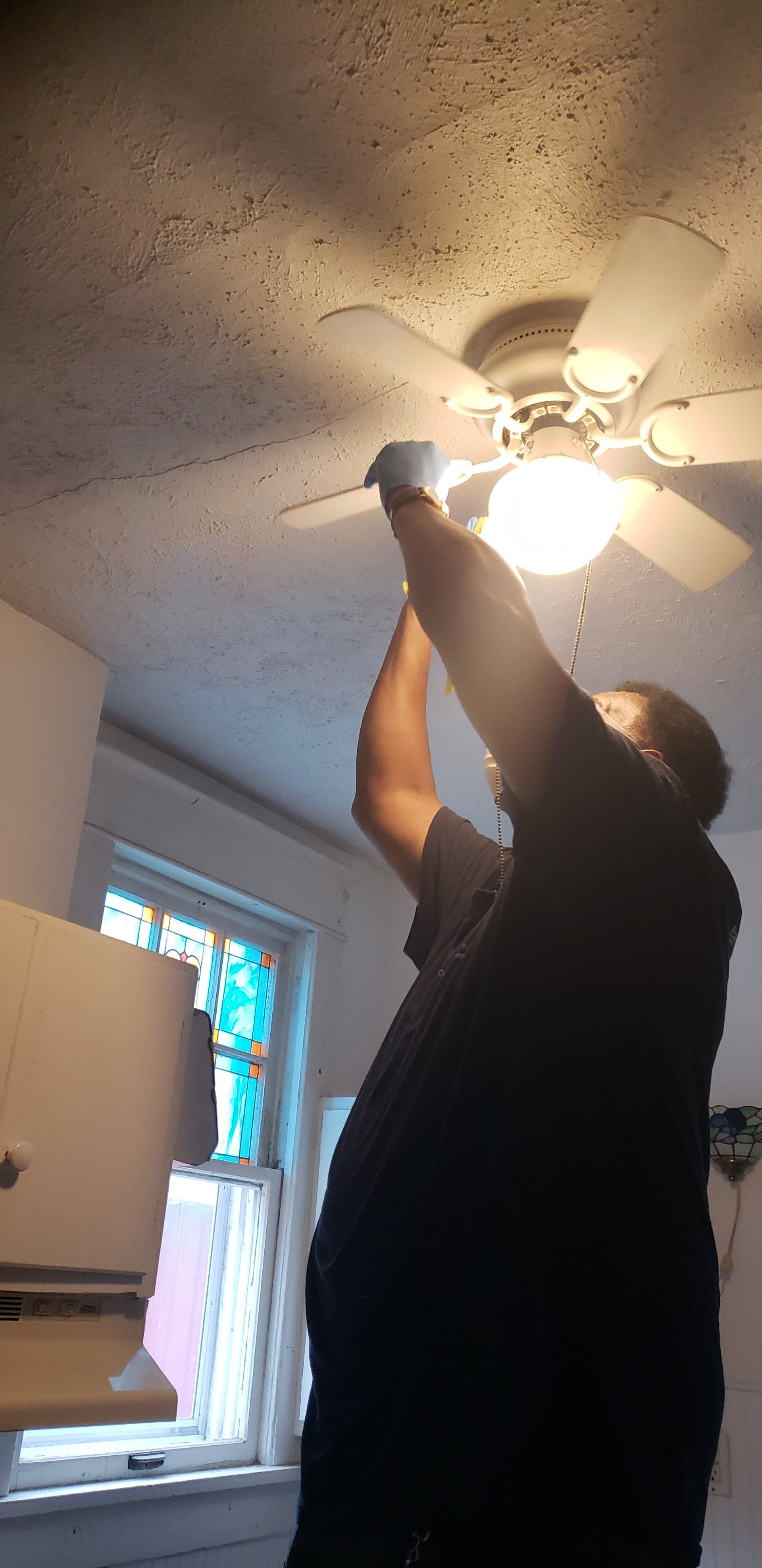 Person reaching up to a ceiling fan. Lit bulb. White walls. Window with colorful stained glass.