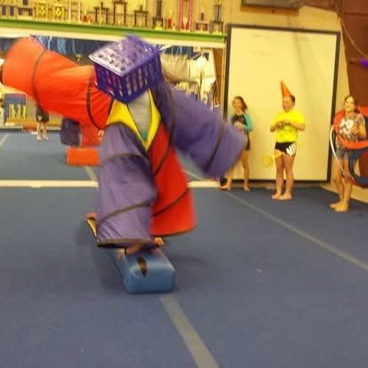 Person in a colorful homemade obstacle costume attempts to navigate on a blue gym floor with others watching.