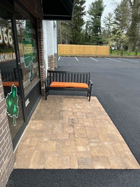Bench with orange cushion in front of store with brick-like pavers; black asphalt; wooden fence in background.