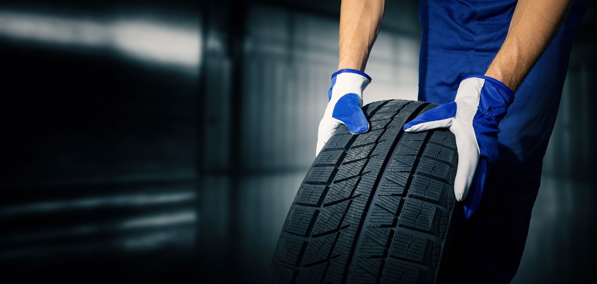 Person in blue uniform and gloves carrying a car tire in an industrial setting.
