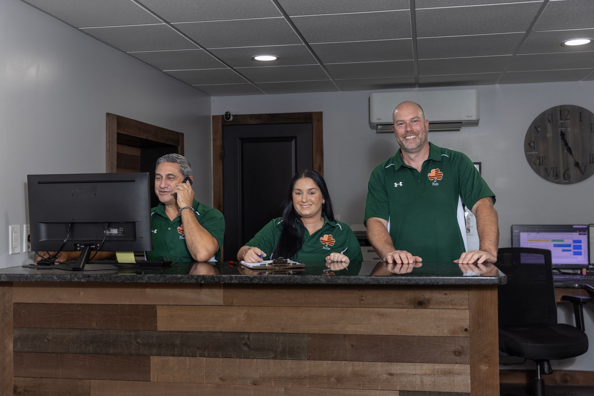 Three people at a front desk; two women and a man. All are wearing green shirts. The desk has a wood front.
