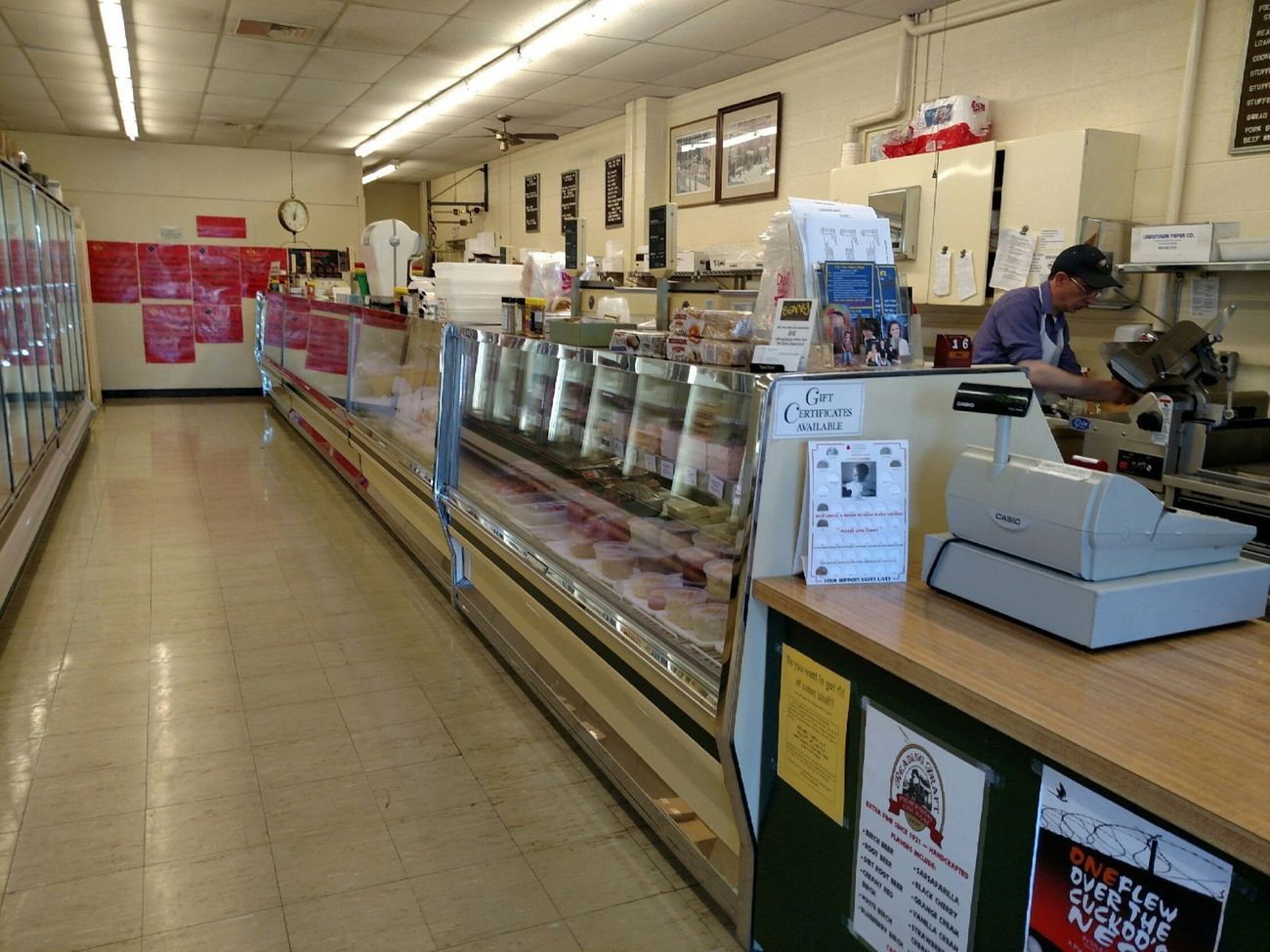 Wide grocery deli counter in a small store, with a cashier at the register and refrigerated display cases.
