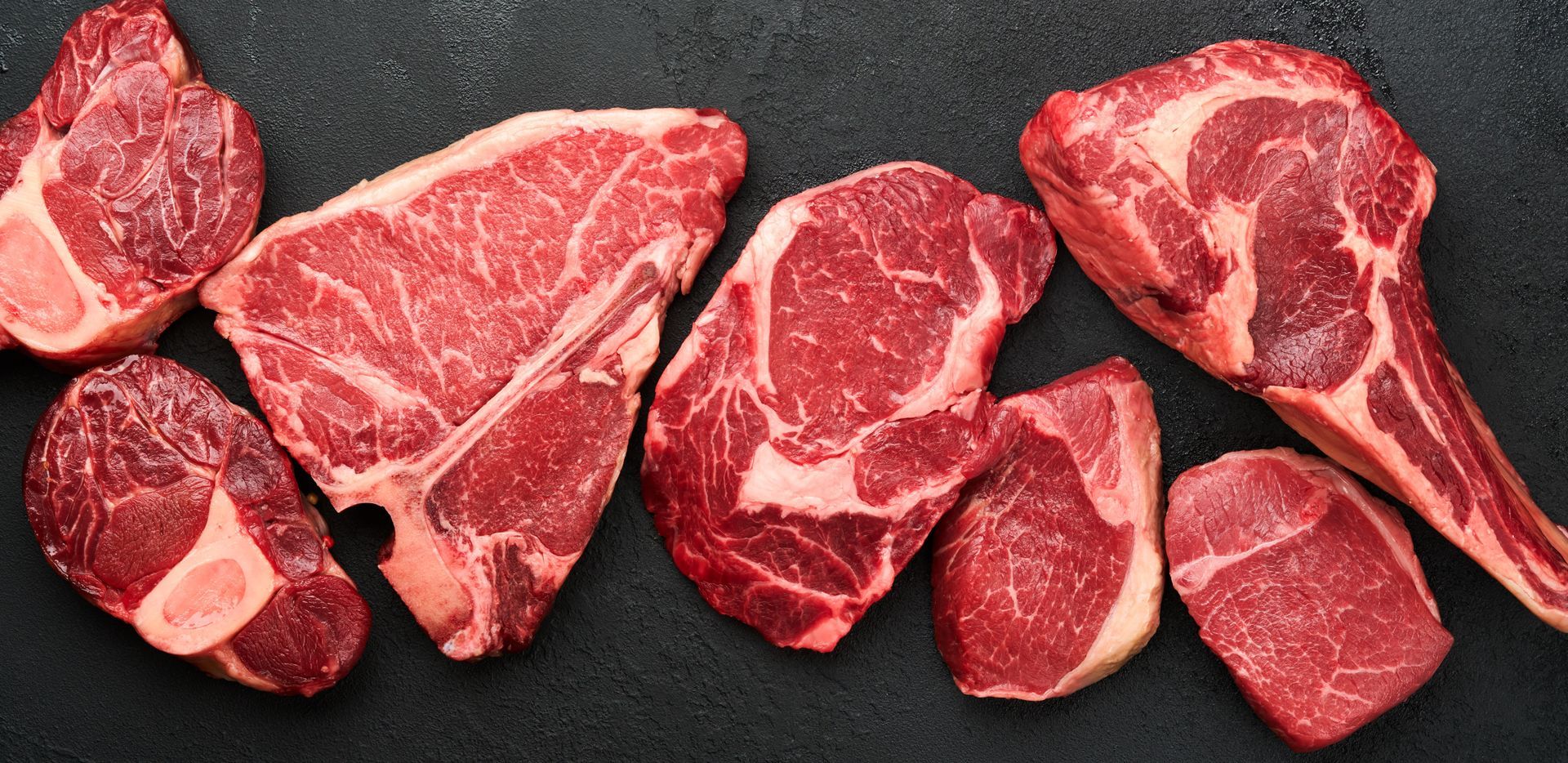 Raw assorted red meat cuts on a dark surface
