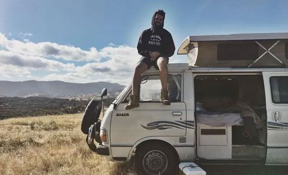 A person sits on the roof of a vintage camper van parked in a grassy field with rolling hills under a blue sky.