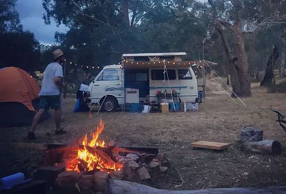 A lit campfire in the foreground with a person walking toward a parked camper van and an orange tent in a wooded area.