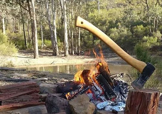 An axe handle rests on a wooden stump over a crackling campfire near a stream in a forest clearing.