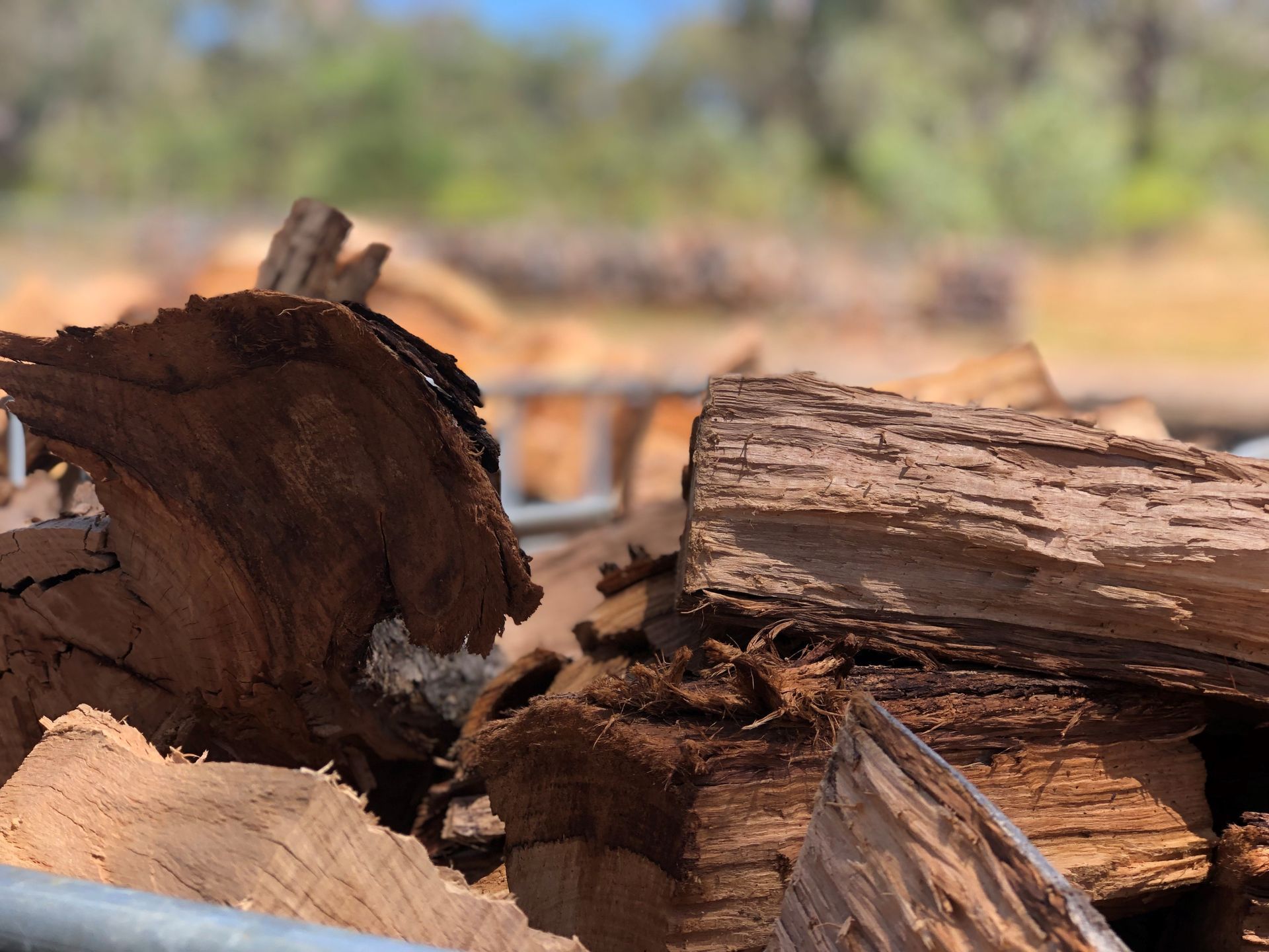 Close-up of stacked, rough-textured pieces of reddish-brown firewood in an outdoor, blurred rural setting.