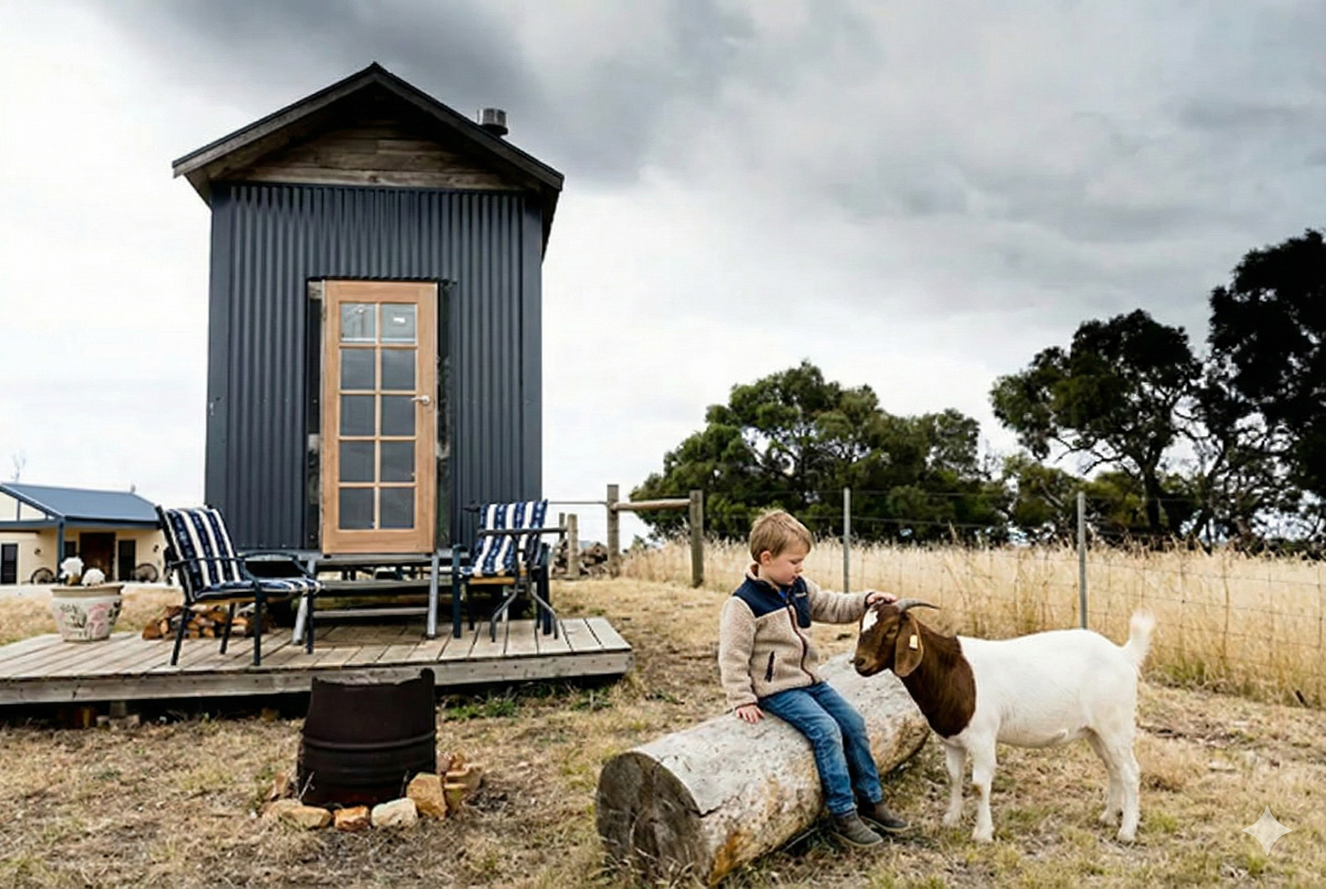 A child sits on a log petting a brown and white goat in front of a dark, narrow tiny house on a grassy rural property.