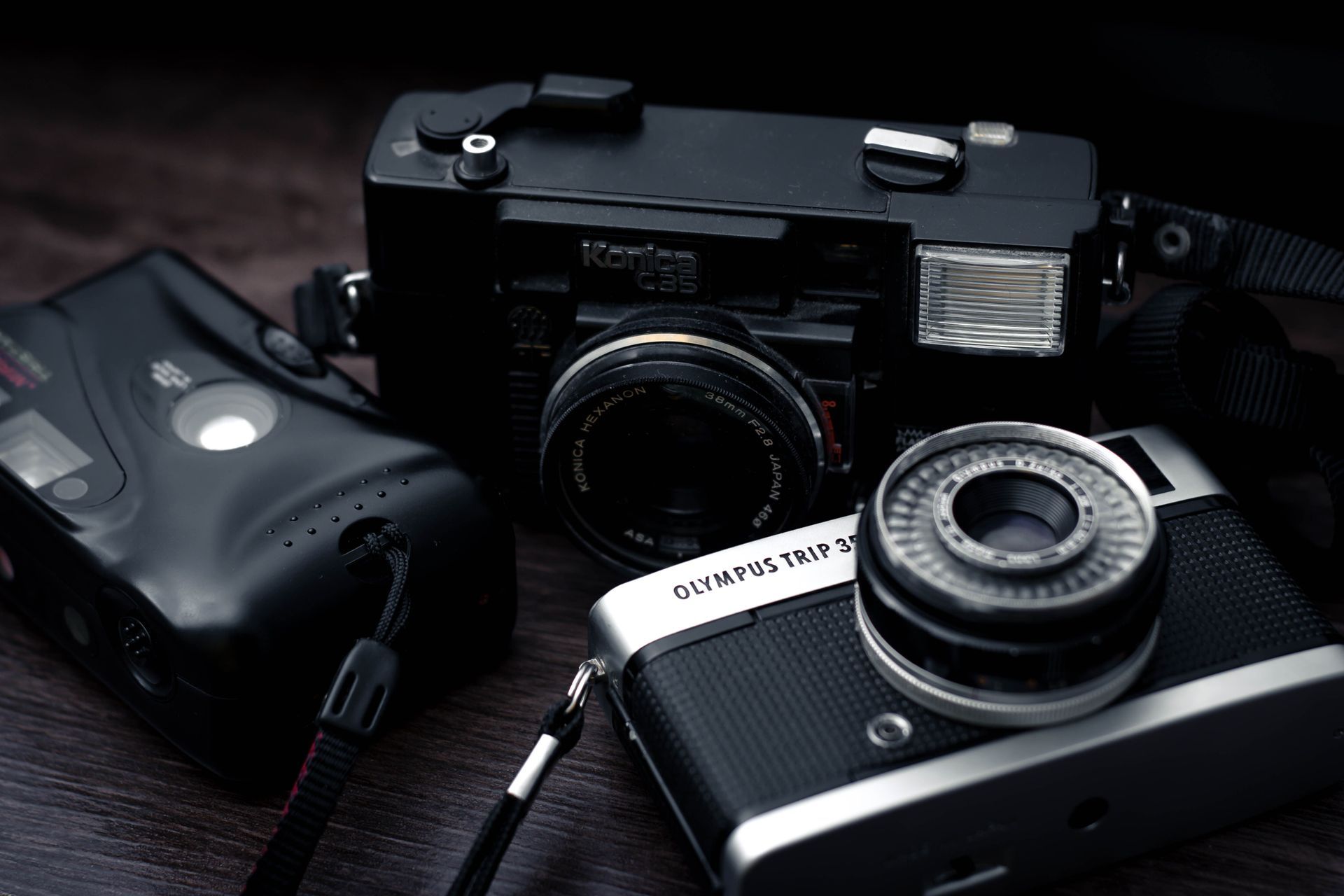 Des appareils photo anciens posés sur une surface sombre, dont un appareil photo argentique noir et un appareil photo compact avec objectif.