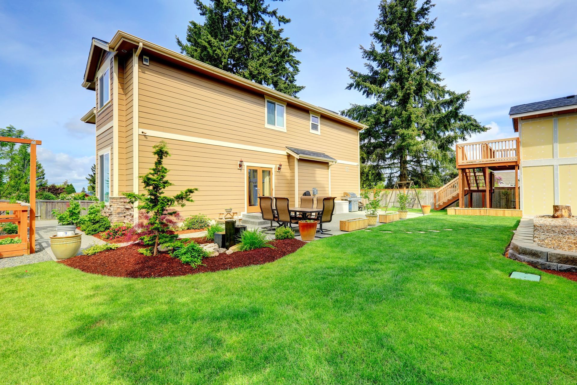 Two-story tan house with a green lawn, patio, and garden, under a bright blue sky.