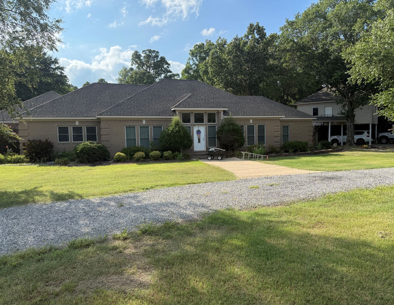 Single-story brick house with a gravel driveway, front porch, and trees under a bright sky.