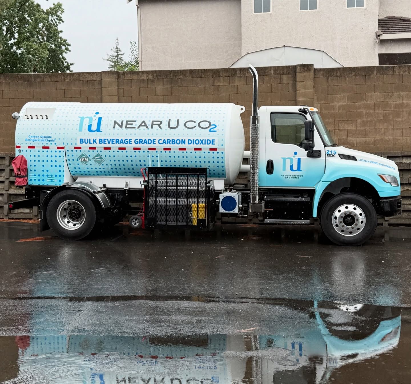 Blue tanker truck labeled “Near U Co” parked on a wet street beside a wall, with its reflection in puddles.