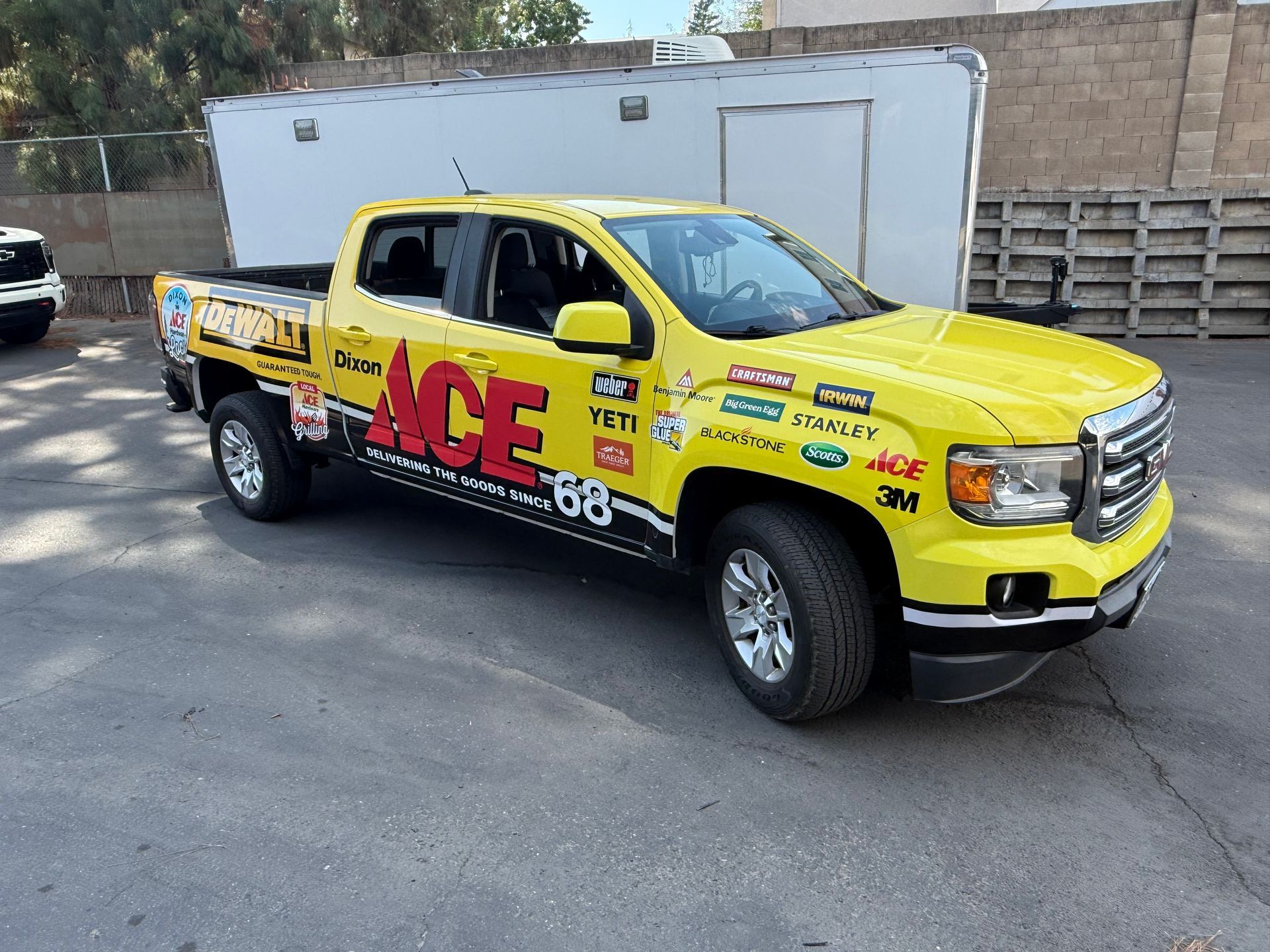 Yellow ACE-branded pickup truck parked on a street beside a white trailer and brick wall.