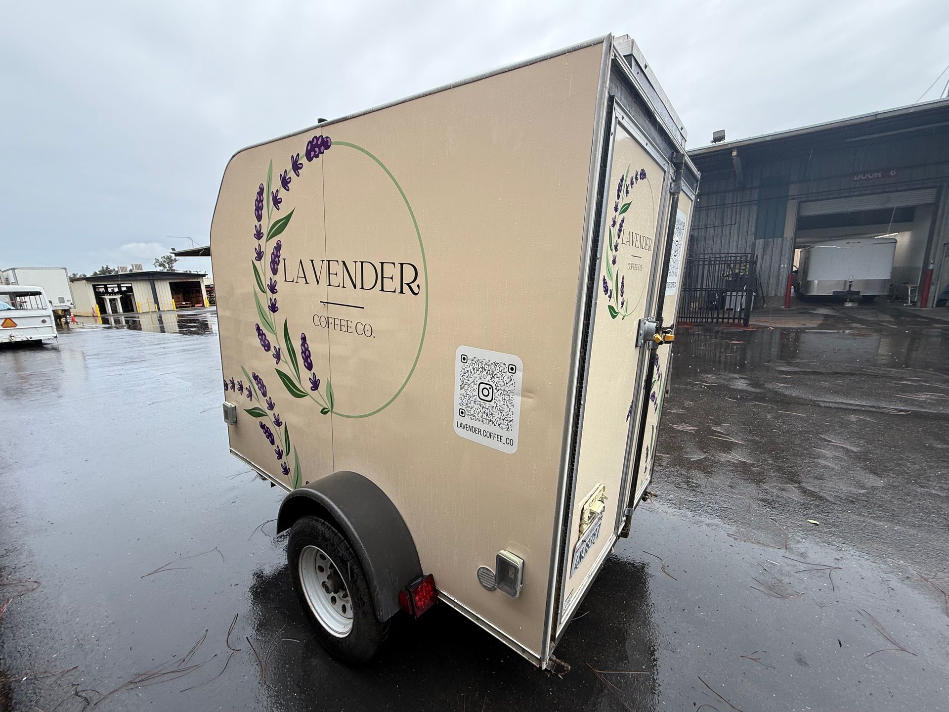 Beige cargo trailer with floral logo parked on a wet lot beside a warehouse