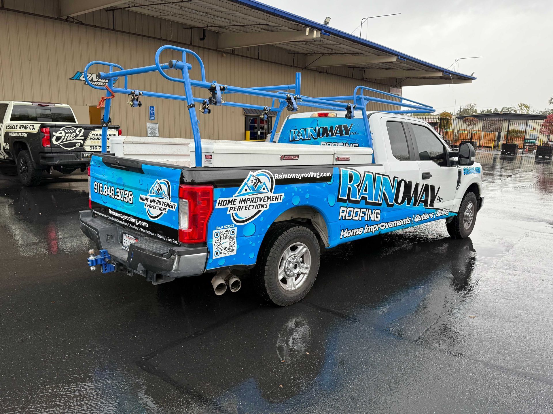 Blue custom pickup truck parked on a wet lot, with racing-style decals and roof rack near a covered building