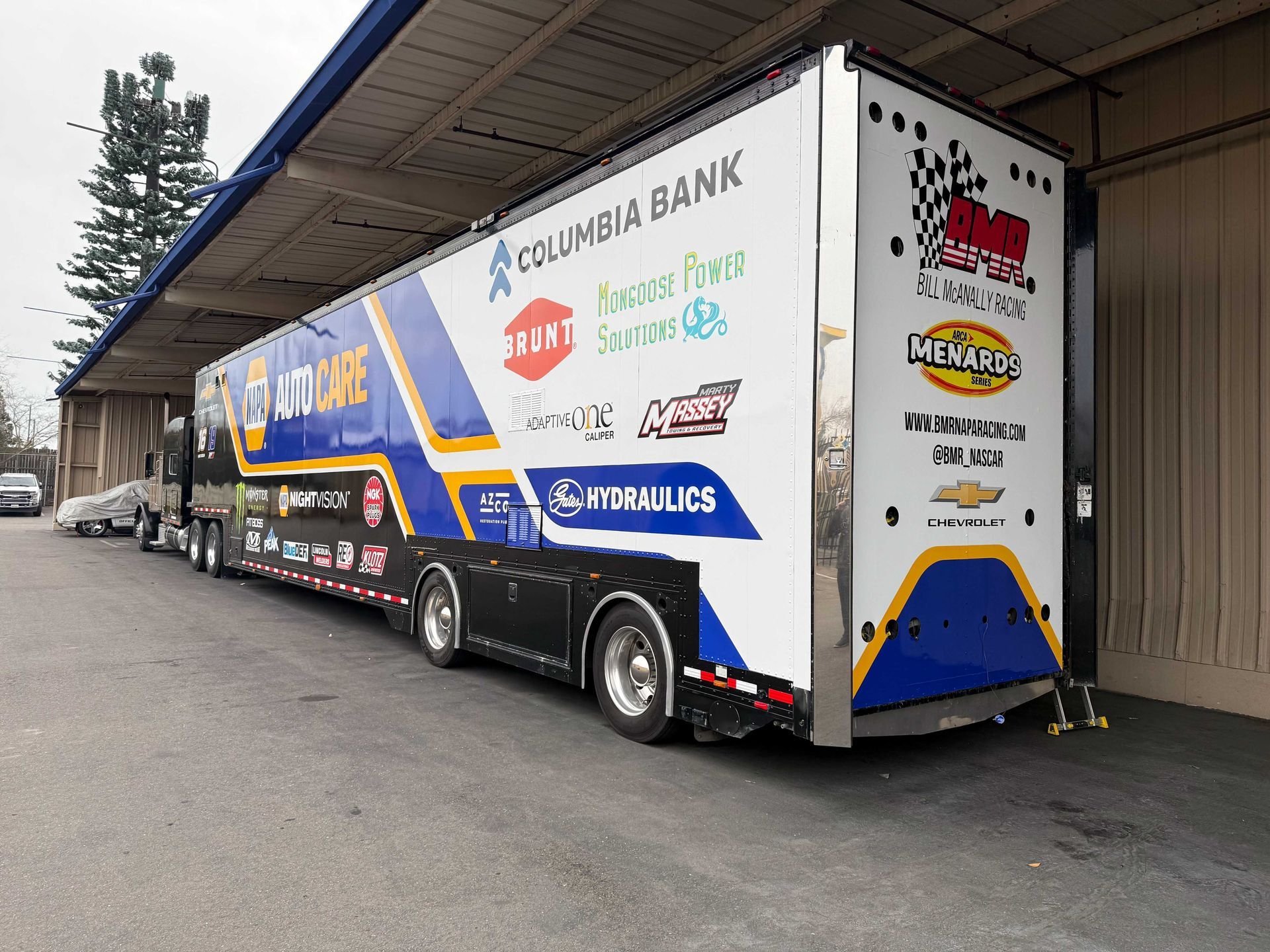 Blue and white race car trailer parked in a warehouse bay, covered with sponsor logos.