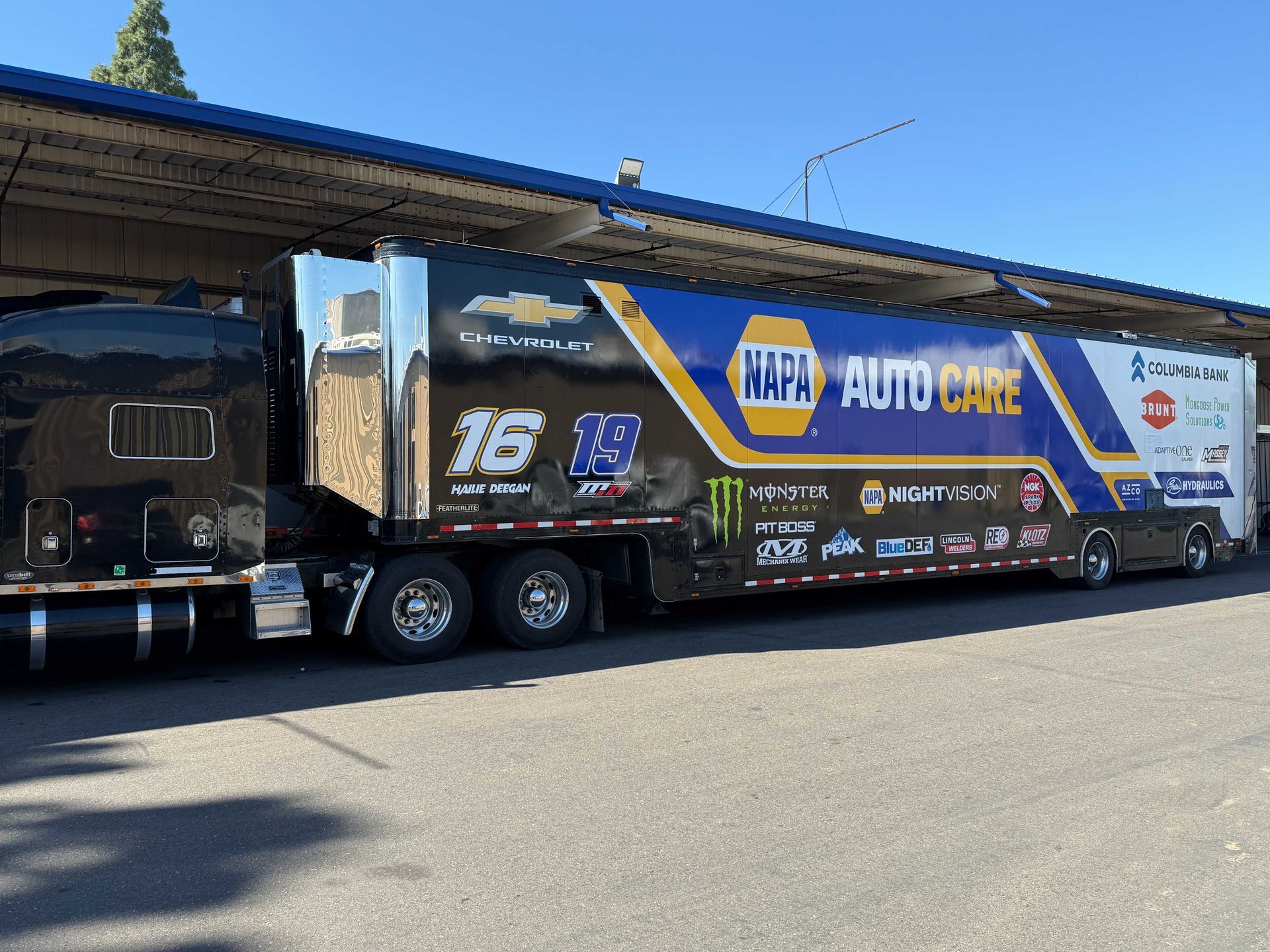 Blue HRT Auto Care racing trailer parked under a canopy, covered with sponsor logos and number 16.