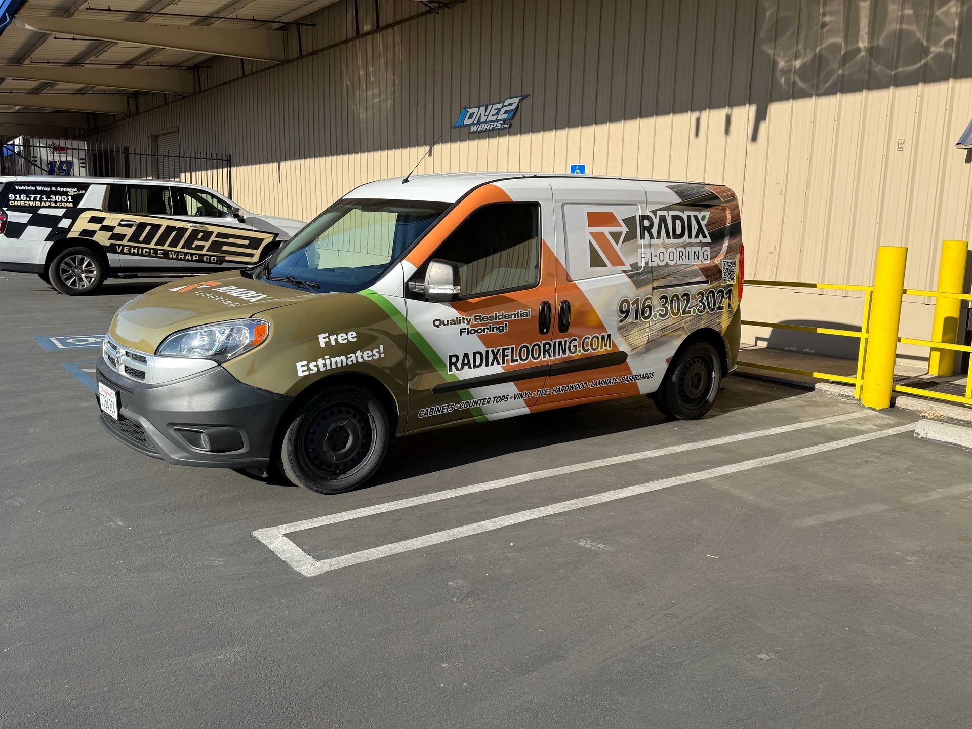 Wrapped silver-and-orange van parked in a loading area beside a building and yellow bollards.