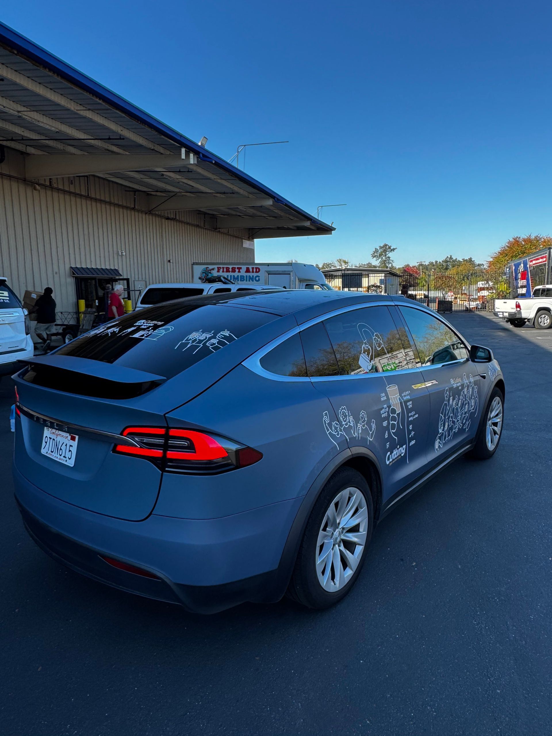 Blue Tesla sedan parked outside a building on a sunny day, with a person loading items nearby.