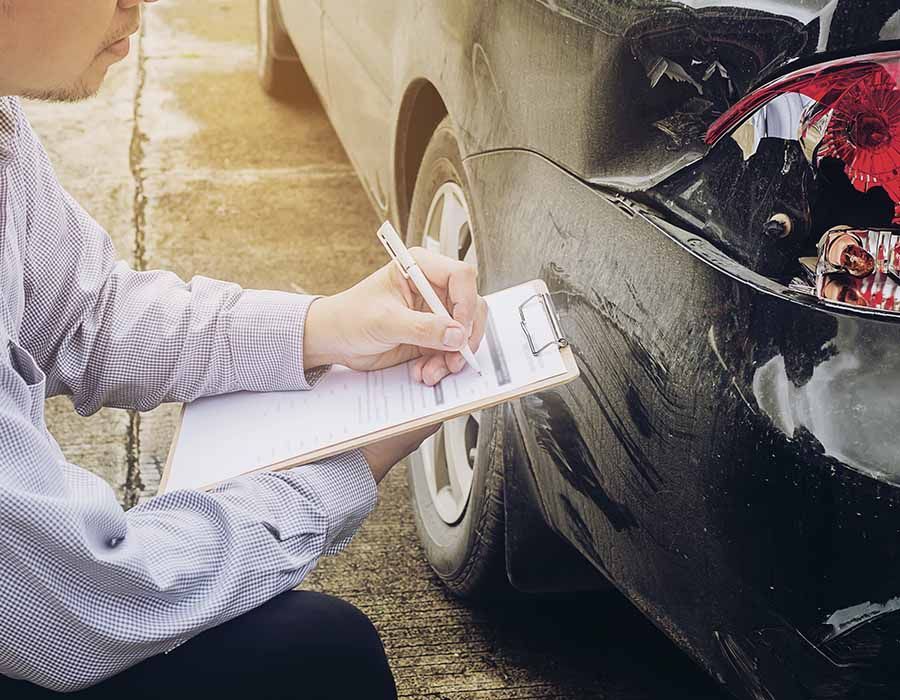Person writing on a clipboard while inspecting damage on a black car's rear bumper.