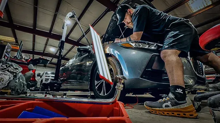 Mechanic crouches beside a silver car in a garage, inspecting the front wheel area.