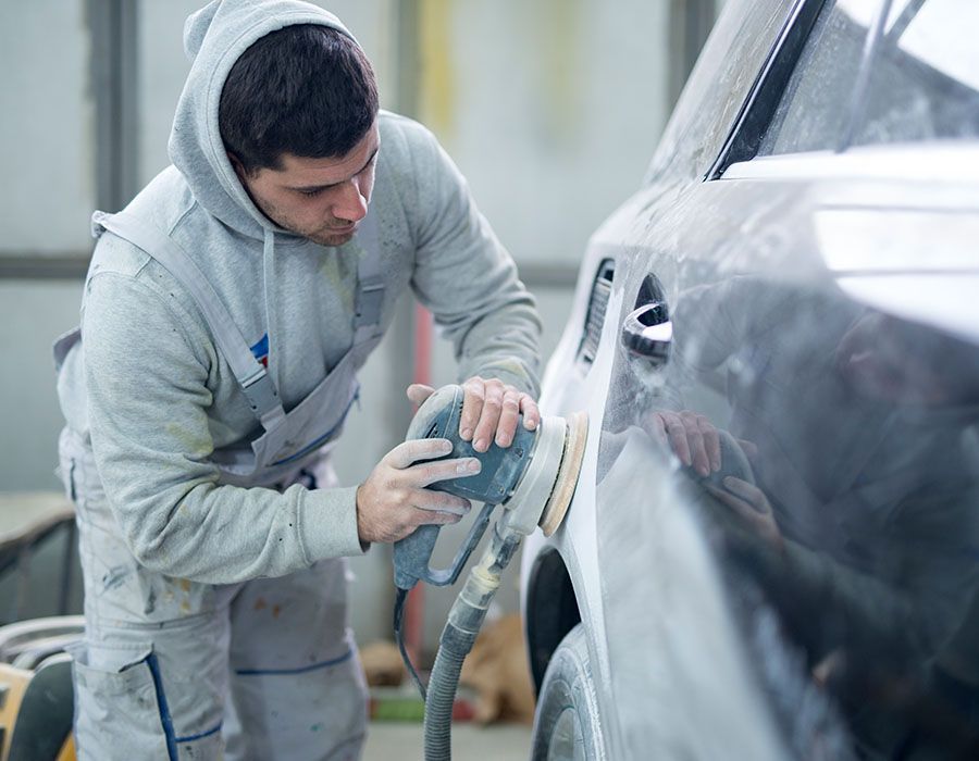 Worker sanding a dark car panel with a power sander in a workshop