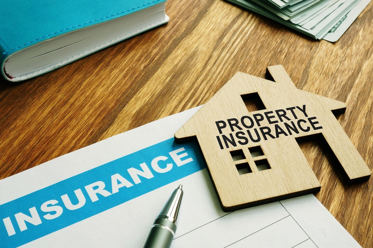 Property insurance documents with house-shaped cutout and pen on a wooden table, with money and a book in the background. Property insurance documents with house-shaped cutout and pen on a wooden table, with money and a book in the background.