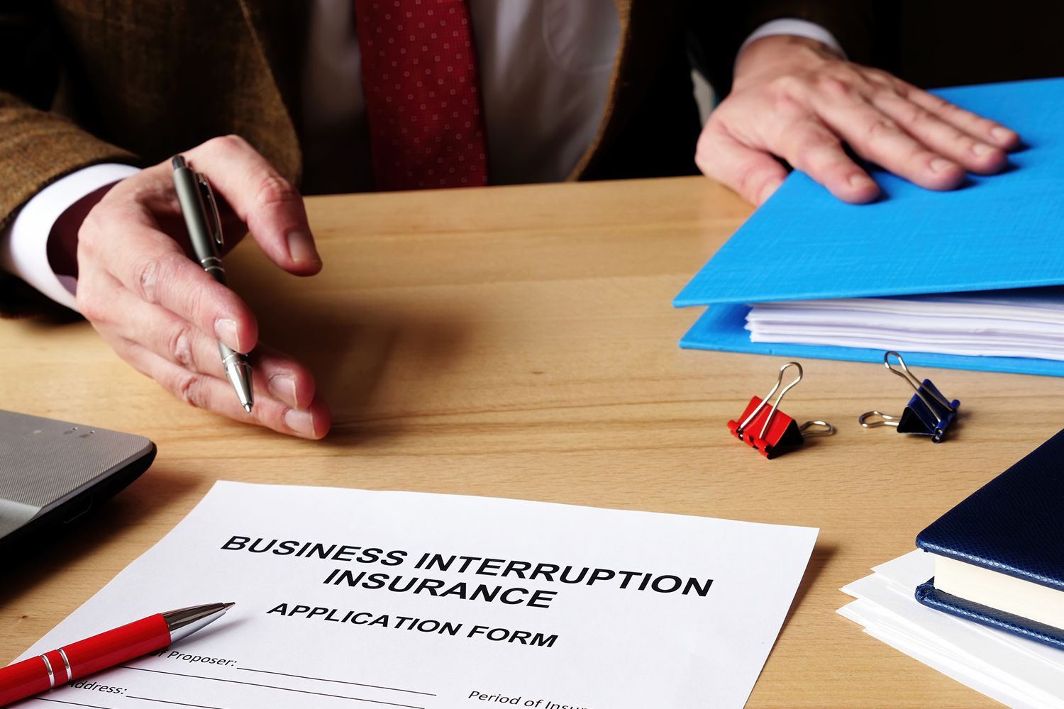 Person with pen over business interruption insurance form on a desk. Blue folder, red pen, and paper clips present. Person with pen over business interruption insurance form on a desk. Blue folder, red pen, and paper clips present.