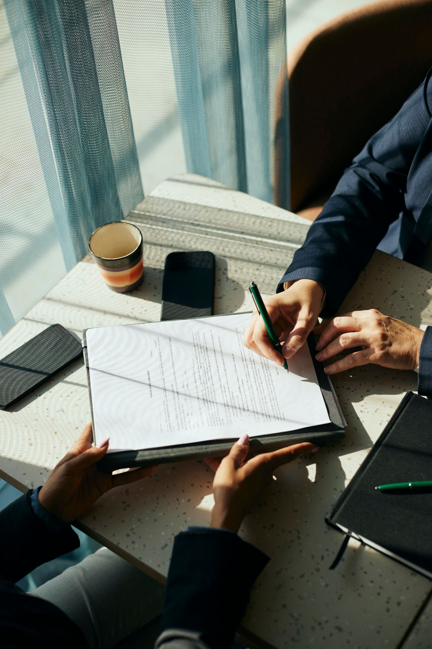Two people in business attire at a table; one signing a document, another holding it. Two people in business attire at a table; one signing a document, another holding it.