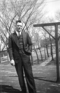 A person in a three-piece suit stands outdoors in a park-like setting with bare trees in the background.