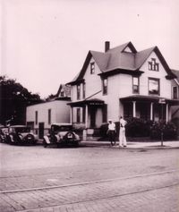A historical sepia photo of a two-story house with a porch, two parked vintage cars, and two people standing on the sidewalk.