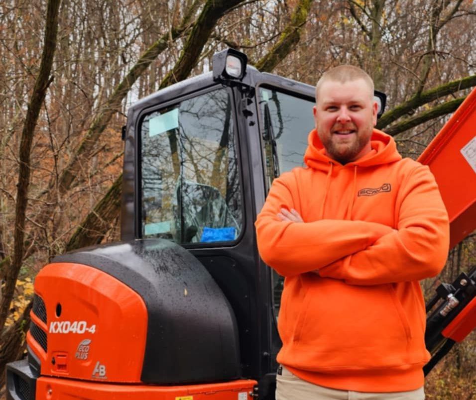 A man in an orange hoodie is standing in front of an orange excavator.