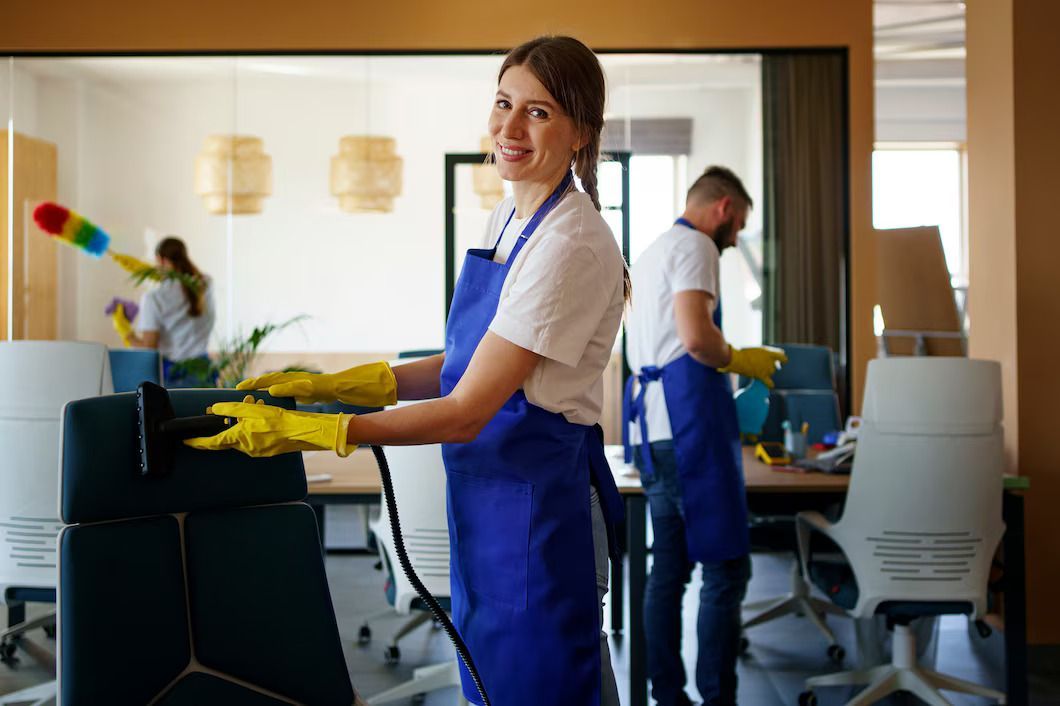 Office cleaners wearing blue aprons and yellow gloves. One woman smiles while wiping. Other cleaners are in the background.