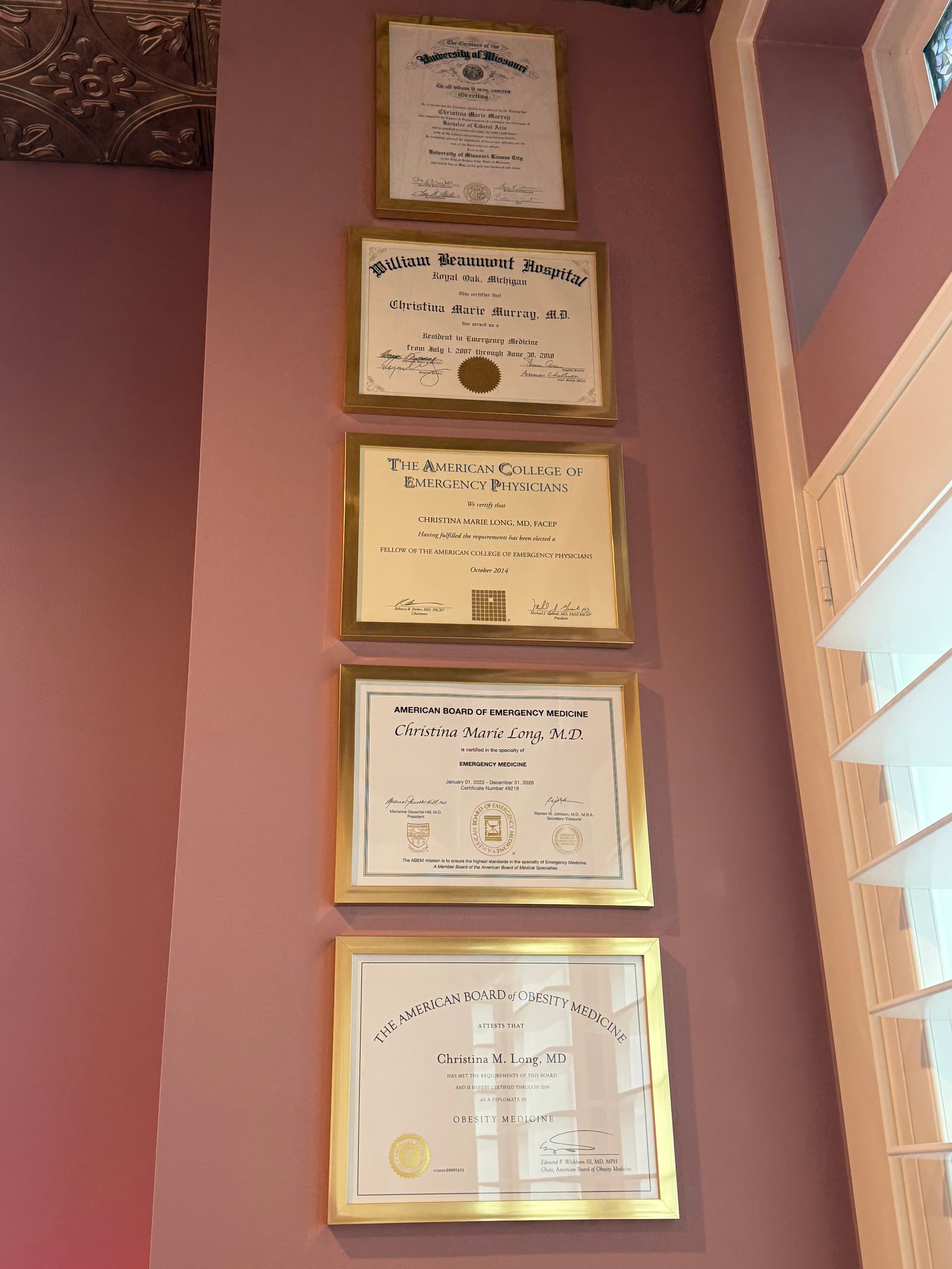 Five framed certificates hanging vertically on a pink wall beside a window with blinds.