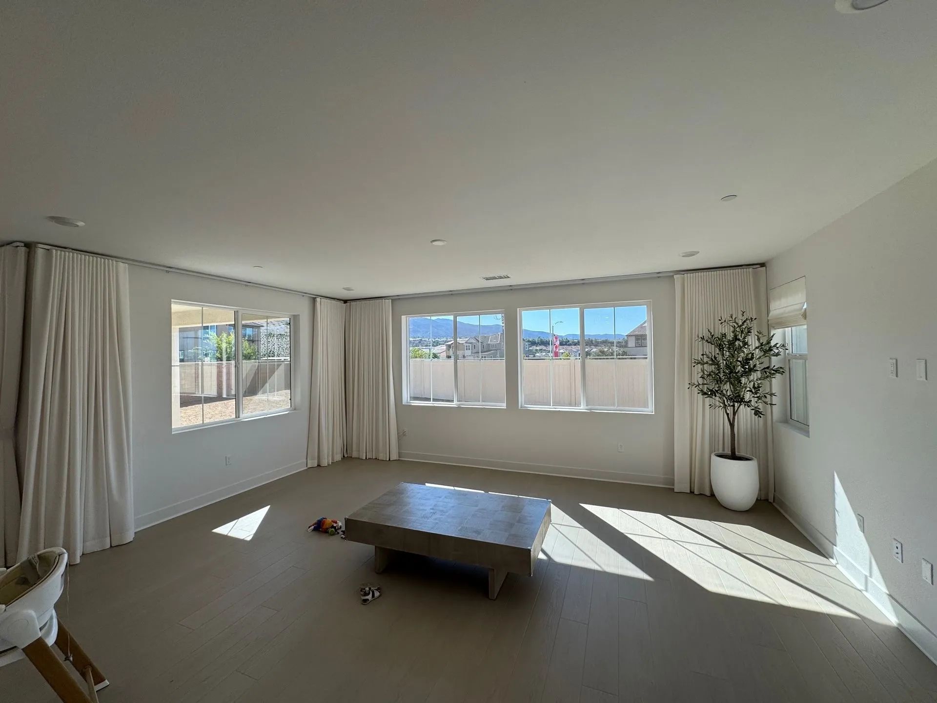 Bright empty living room with large windows, white curtains, and a low coffee table on carpeted floor.