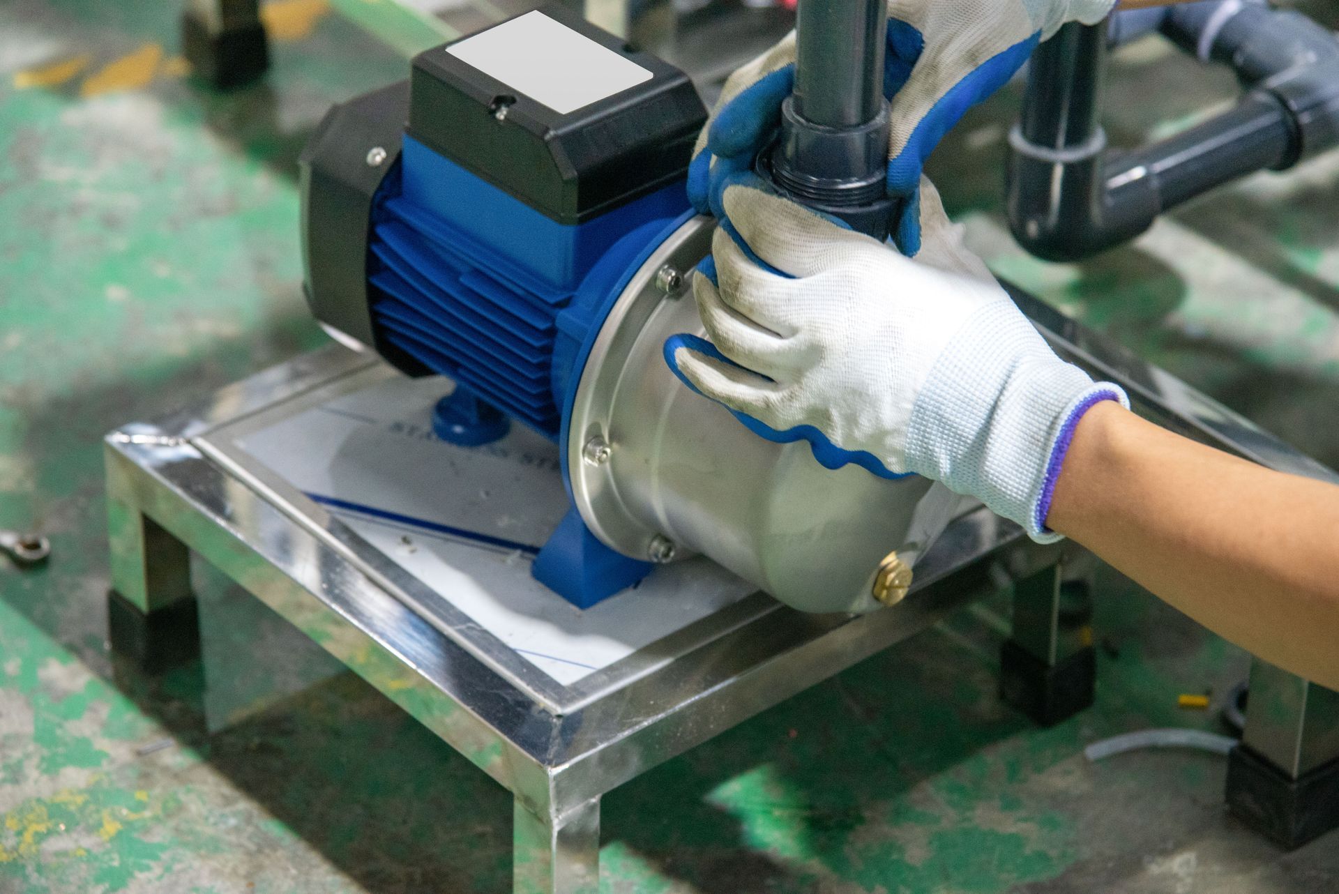 A person wearing work gloves installs plumbing onto a blue industrial water pump mounted on a stainless steel stand. A person wearing work gloves installs plumbing onto a blue industrial water pump mounted on a stainless steel stand.
