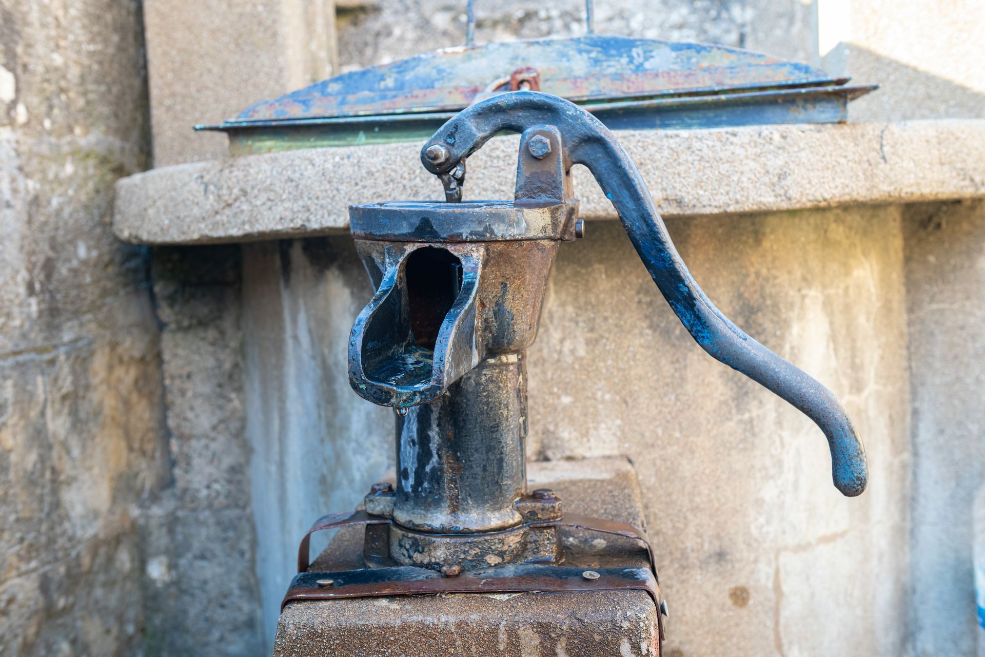 An antique black cast-iron hand water pump mounted on a stone base in front of a concrete well structure.