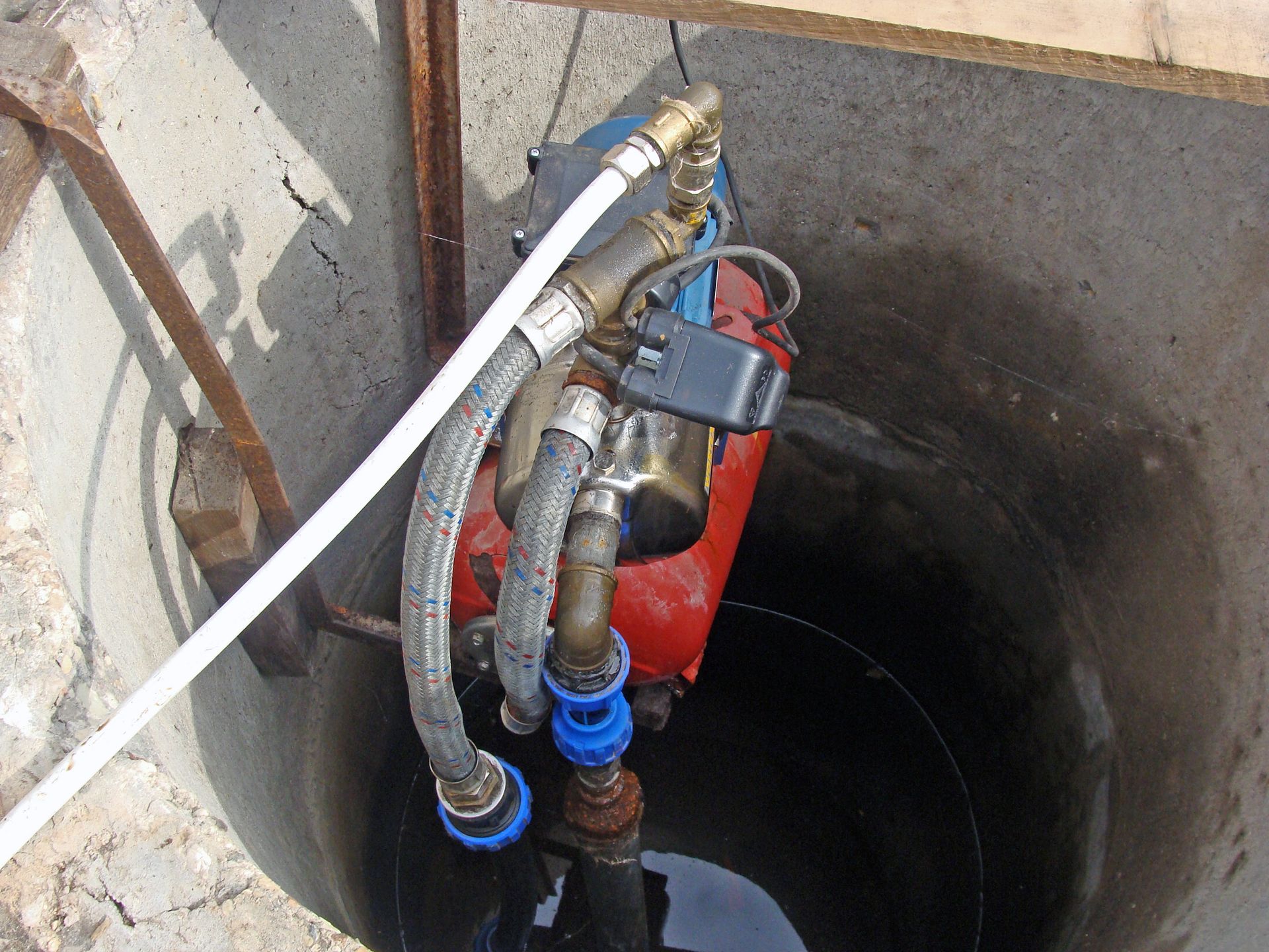 A small, red water pump installed inside a concrete well, connected with blue fittings and various hoses. A small, red water pump installed inside a concrete well, connected with blue fittings and various hoses.