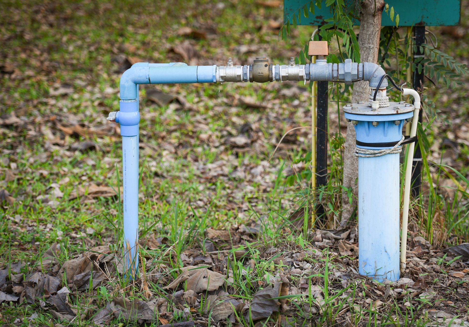 A blue PVC pipe assembly with a metal valve connects a vertical well pipe to a ground-level pipe in a grassy, leaf-covered area. A blue PVC pipe assembly with a metal valve connects a vertical well pipe to a ground-level pipe in a grassy, leaf-covered area.