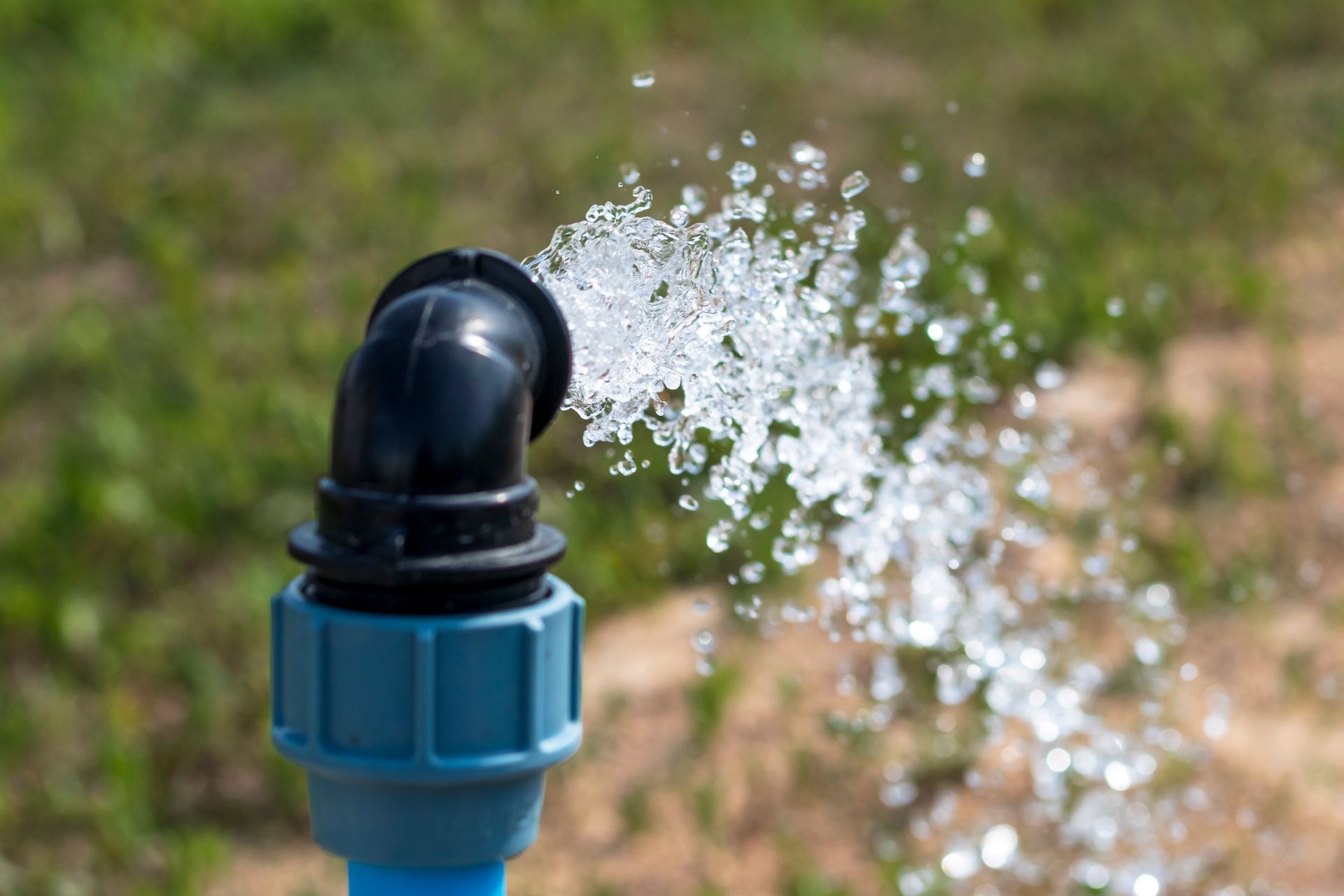 Water sprays from a black angled pipe fitting with a blue connector against a blurry green and brown natural background.
