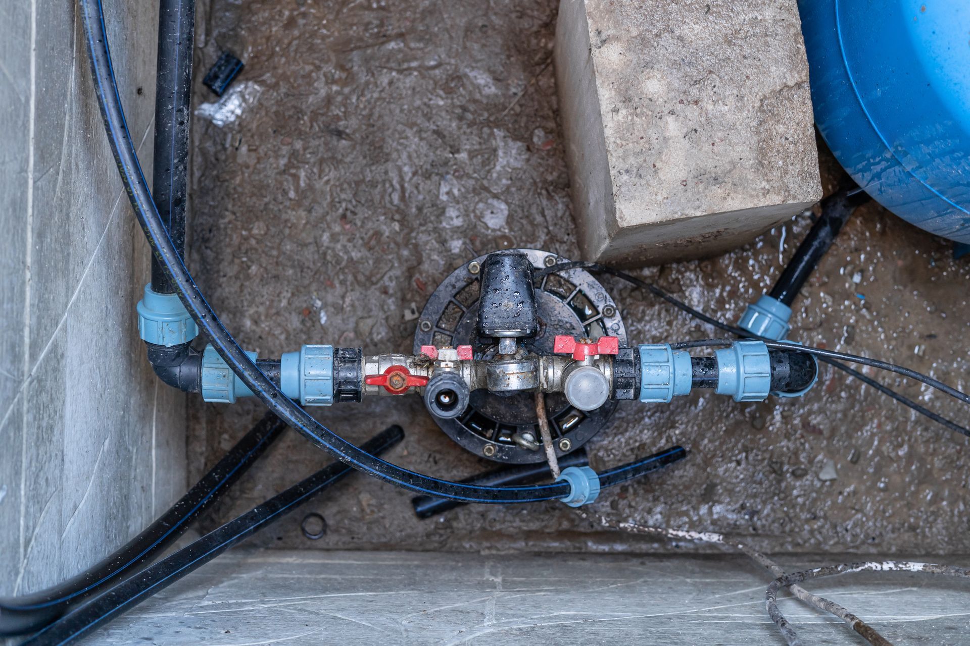 An irrigation system manifold with black pipes and blue connectors mounted on a dirt ground surface next to a concrete block. An irrigation system manifold with black pipes and blue connectors mounted on a dirt ground surface next to a concrete block.