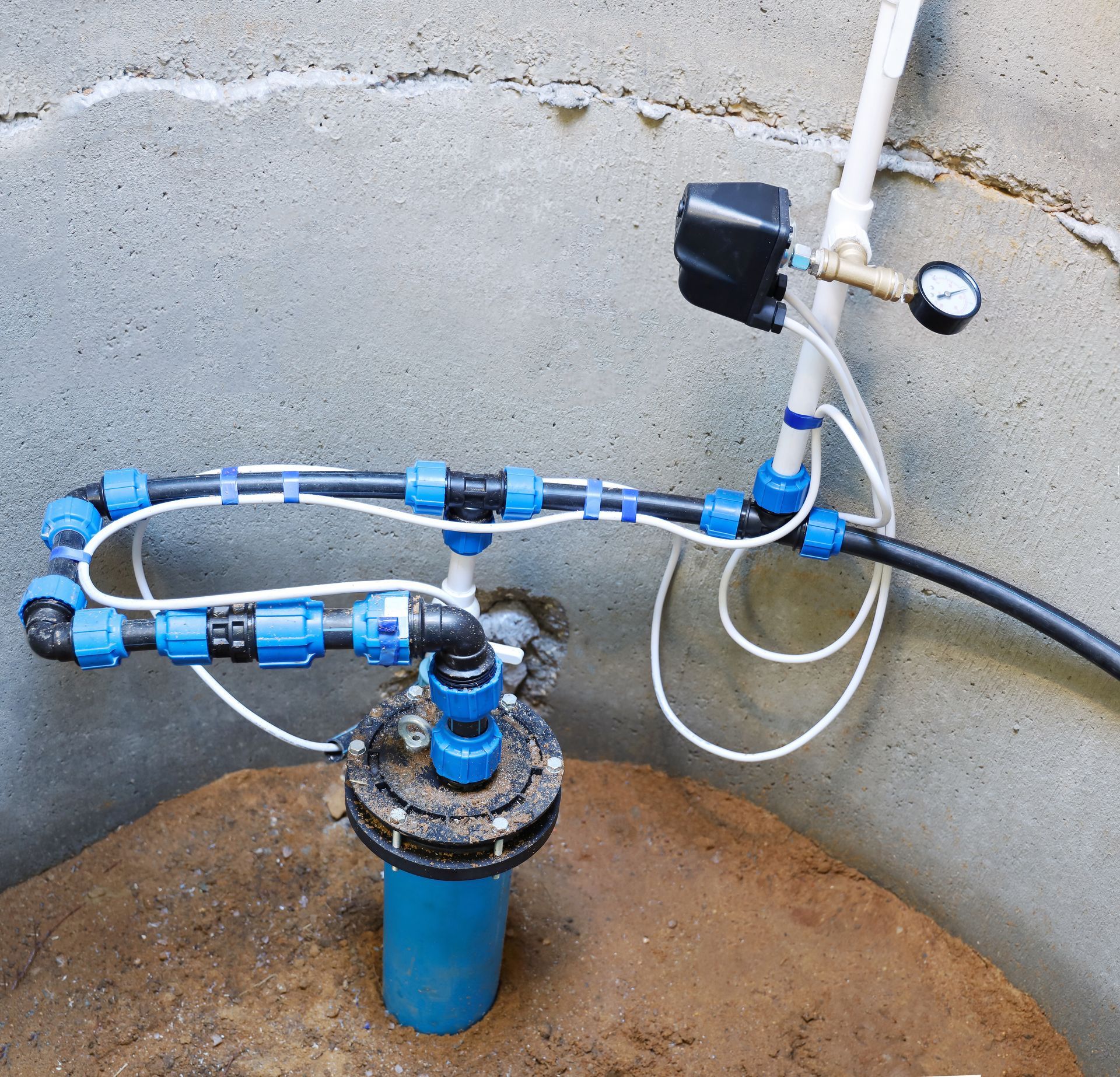 Water well pump assembly with blue plastic pipes, a pressure gauge, and electrical wiring inside a concrete enclosure. Water well pump assembly with blue plastic pipes, a pressure gauge, and electrical wiring inside a concrete enclosure.