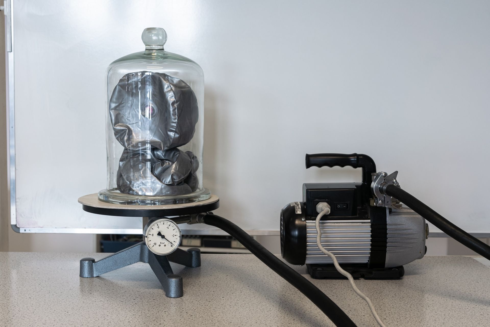 A vacuum pump connected to a glass bell jar containing a partially crushed metallic object on a table. A vacuum pump connected to a glass bell jar containing a partially crushed metallic object on a table.
