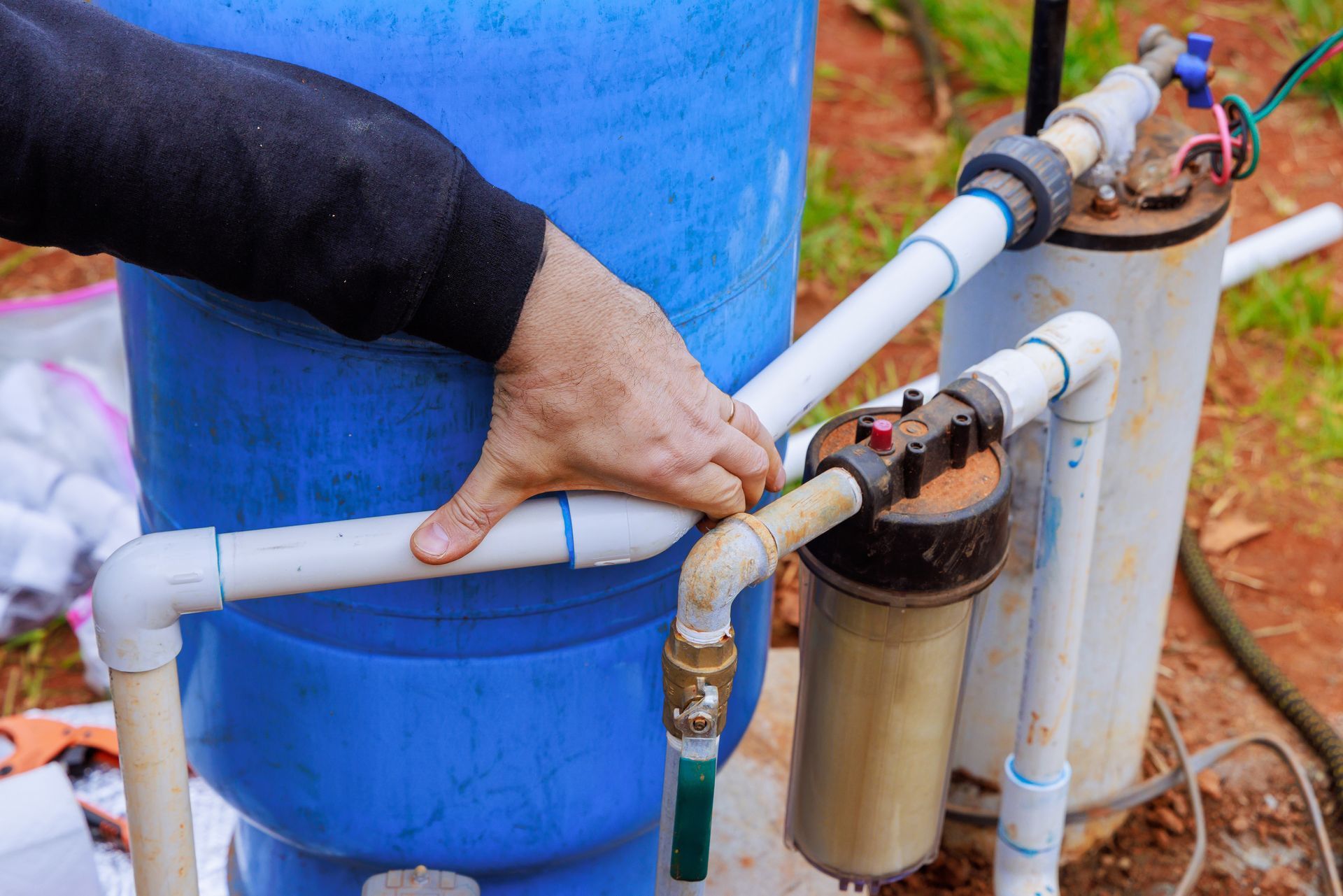 A person's hand touches white PVC pipes connected to a blue water tank and a vertical cylindrical filter outdoors. A person's hand touches white PVC pipes connected to a blue water tank and a vertical cylindrical filter outdoors.