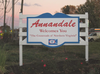 A white welcome sign for Annandale, Virginia, featuring red text and a small emblem, set outdoors at dusk. A white welcome sign for Annandale, Virginia, featuring red text and a small emblem, set outdoors at dusk.