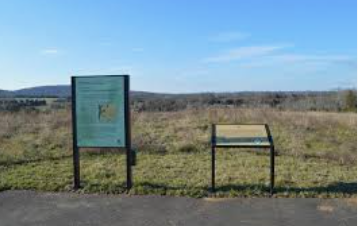Trail sign and bench beside a paved path in a grassy open field under a blue sky Trail sign and bench beside a paved path in a grassy open field under a blue sky