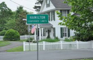 Hamilton corporate limits sign in front of a white house with a picket fence and American flag Hamilton corporate limits sign in front of a white house with a picket fence and American flag