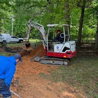 Man operating small excavator digging in yard, another man observing. White truck in background.