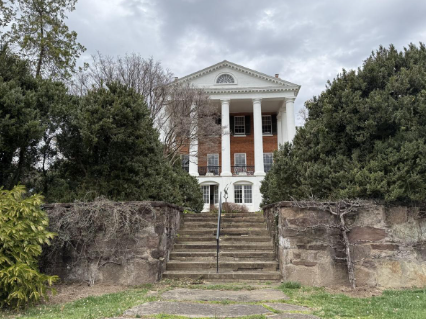 A large, white-pillared historic brick mansion viewed from the bottom of stone steps surrounded by evergreen bushes.